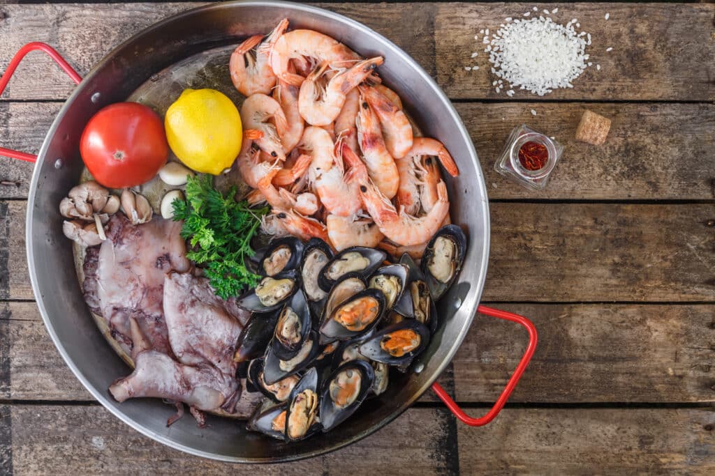 Raw seafood products in paella pan on a wooden table, top view